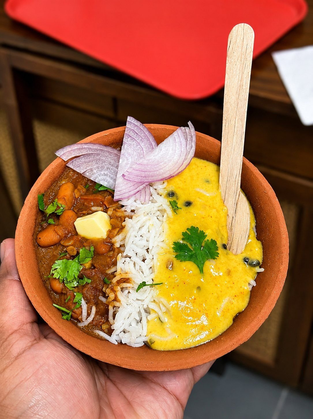 A clay bowl of rajma, rice and kadhi held out in one hand, finished with butter, sliced onion and coriander
