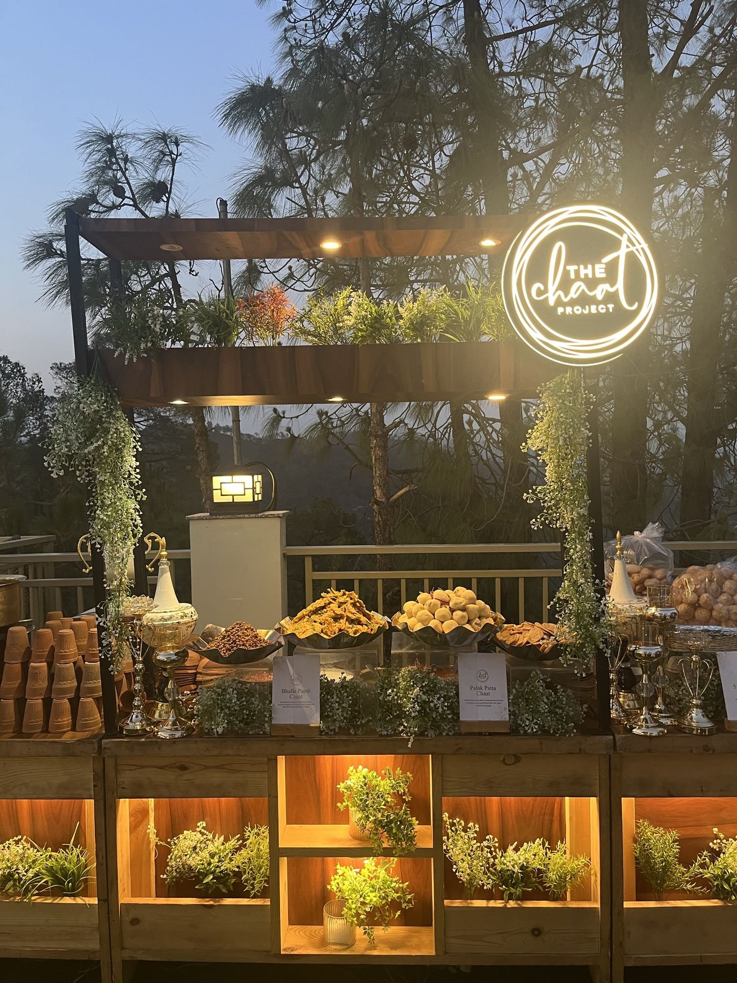 A live counter set up outdoors at dusk, golgappa and papdi in glass jars under the lit Chaat Project sign